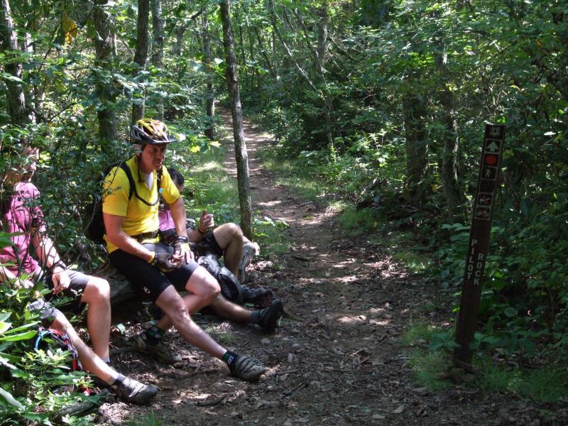 Three mountain bikers resting on a log beside a wooded trail. The trail is surrounded by greenery, and a signpost indicating the Pilot Rock trail is visible nearby. The bikers are dressed in cycling gear, with one wearing a yellow shirt and helmet. Laurel Mountain mountain bike trail.