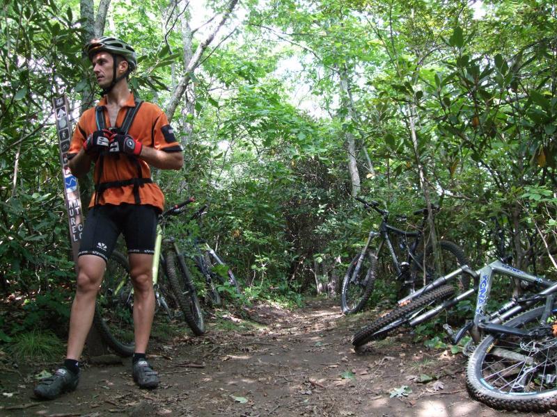 A mountain biker wearing an orange jersey and black shorts stands on a narrow dirt trail surrounded by dense greenery. Several mountain bikes are parked nearby, partially obscured by bushes. A trail sign is visible in the background, indicating the path. The bright sunlight filters through the trees, creating a vibrant outdoor scene. Laurel Mountain mountain bike trail.