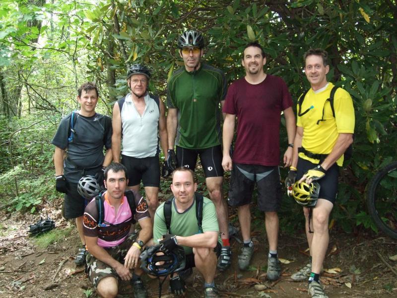A group of seven mountain bikers posing together in a wooded area, with lush greenery in the background. They are wearing cycling gear and helmets, with some seated and others standing. The group appears to be enjoying a break from their biking activity. Laurel Mountain mountain bike trail.