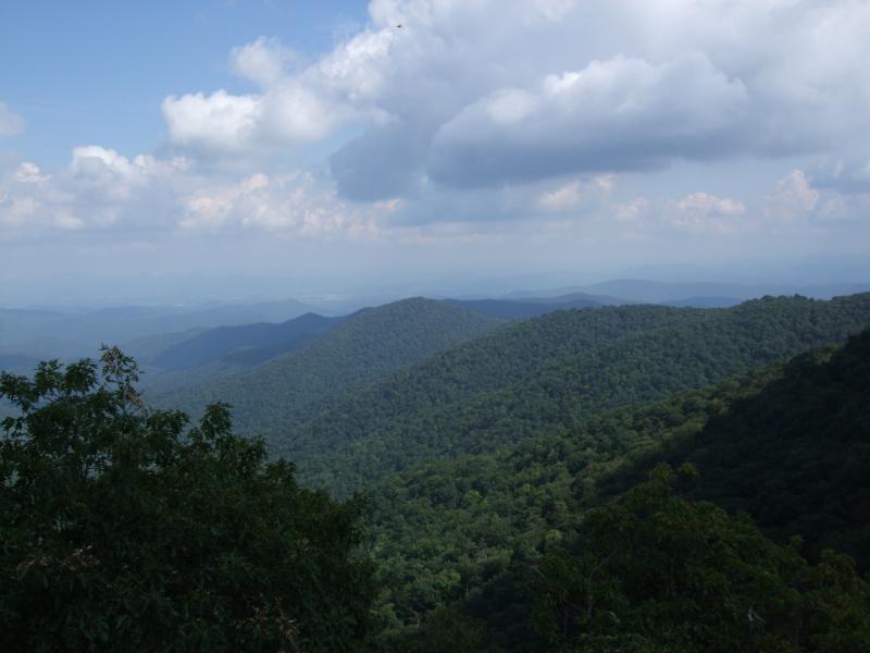 A scenic view of rolling mountains covered in lush green forests under a partly cloudy sky. The landscape features layers of hills extending into the distance, showcasing the natural beauty of the terrain. Laurel Mountain mountain bike trail.