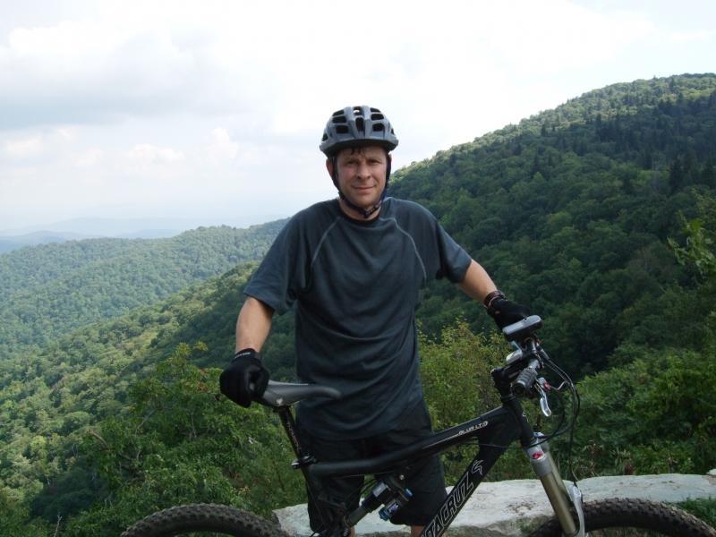 A person in a helmet and biking gloves stands beside a mountain bike on a scenic overlook, with lush green mountains in the background under a partly cloudy sky. Laurel Mountain mountain bike trail.