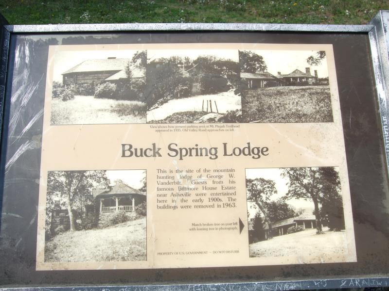 A historical plaque titled "Buck Spring Lodge" that features several black and white photographs of the former hunting lodge of George W. Vanderbilt. The text explains the lodge's significance, stating it was a site where guests from the Biltmore Estate near Asheville were entertained in the early 1900s, and mentions that the buildings were removed in 1963. The plaque has a protective cover and is set against a grassy background. Laurel Mountain mountain bike trail.