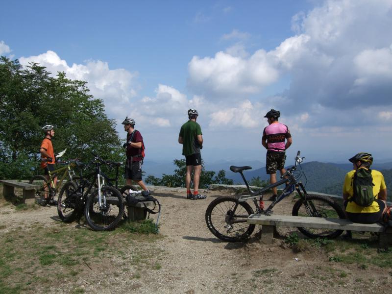 Five mountain bikers are standing and sitting on a rocky overlook, enjoying a scenic view of rolling hills and cloudy skies. They are wearing helmets and colorful cycling attire, with their bikes parked nearby. Some are gazing at the landscape, while one person sits on a wooden bench, facing the view. The area is surrounded by trees and nature. Laurel Mountain mountain bike trail.