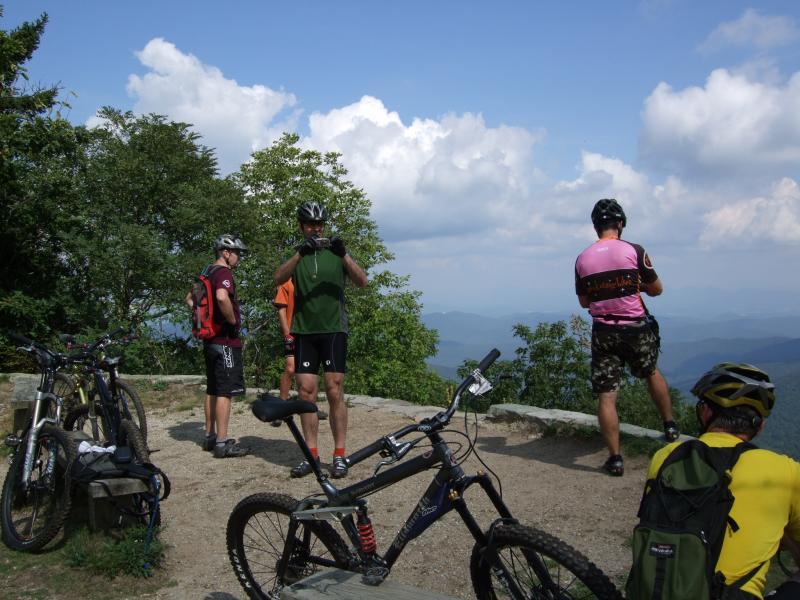 Four mountain bikers are gathered at a scenic viewpoint, overlooking a landscape of rolling hills and mountains under a partly cloudy sky. Various bicycles are parked nearby, and the cyclists are wearing helmets and athletic clothing, engaged in discussion and taking in the view. Laurel Mountain mountain bike trail.
