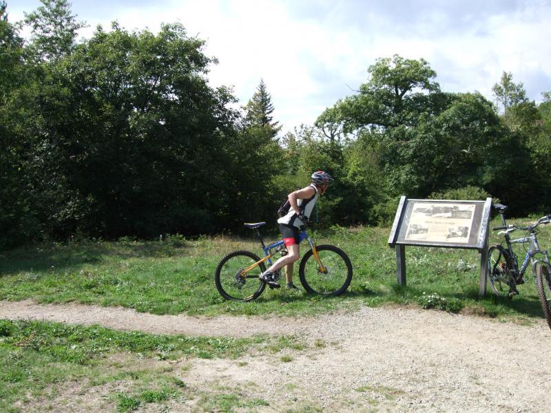 A person in cycling gear adjusting their mountain bike next to a trail sign, surrounded by trees and greenery. Two additional bikes are parked nearby, and the scene is set on a sunny day. Laurel Mountain mountain bike trail.