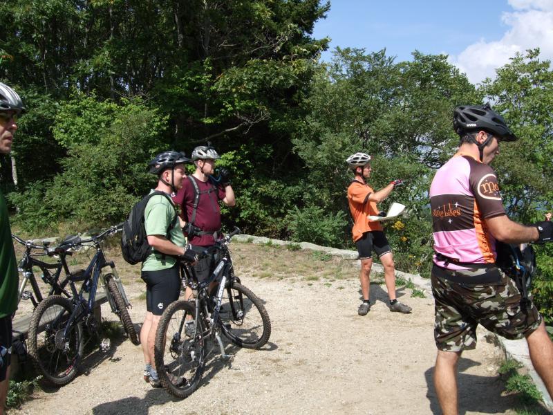 A group of five mountain bikers, wearing helmets and cycling gear, stands near their bikes on a dirt trail surrounded by greenery. One person is pointing and discussing a map, while the others listen attentively. The scene captures a moment of planning or instruction during a biking excursion. Laurel Mountain mountain bike trail.