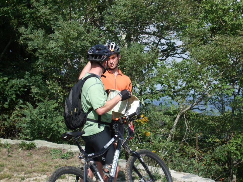 Two cyclists are standing in a wooded area, discussing a map. One cyclist, wearing a green shirt and a helmet, is facing slightly away while holding the map. The other cyclist, dressed in an orange shirt, is looking at him attentively. Bicycles are parked nearby, and the background features greenery and distant trees. Laurel Mountain mountain bike trail.