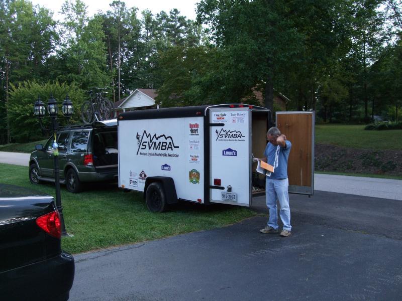 A man stands next to an open trailer parked in a driveway, looking at papers in his hand. The trailer, branded with "SVMBA" and various logos, is surrounded by trees and a suburban home in the background. A bicycle is mounted on the roof of the vehicle parked nearby. Laurel Mountain mountain bike trail.
