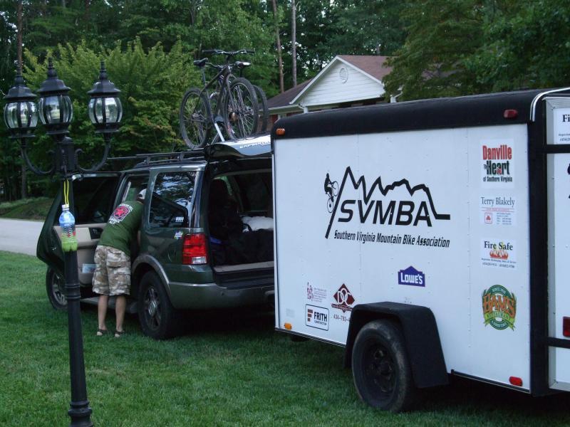 A green SUV with a mountain bike rack on top is parked next to a white trailer marked with "Southern Virginia Mountain Bike Association." A person in a green shirt and camo shorts is seen reaching into the back of the SUV. In the background, there are trees and a residential home. A decorative street lamp stands in the foreground. Laurel Mountain mountain bike trail.
