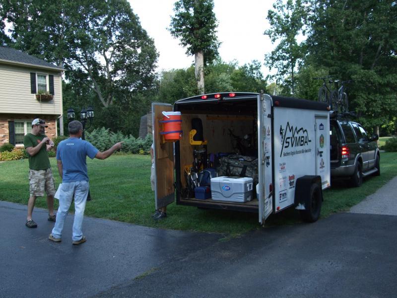 Two men are standing outside in a residential area, discussing while facing a trailer that is parked nearby. The trailer, which is open at the back, is filled with various equipment, including bicycles and a cooler. Trees and a house are visible in the background, suggesting an outdoor activity setting. Laurel Mountain mountain bike trail.