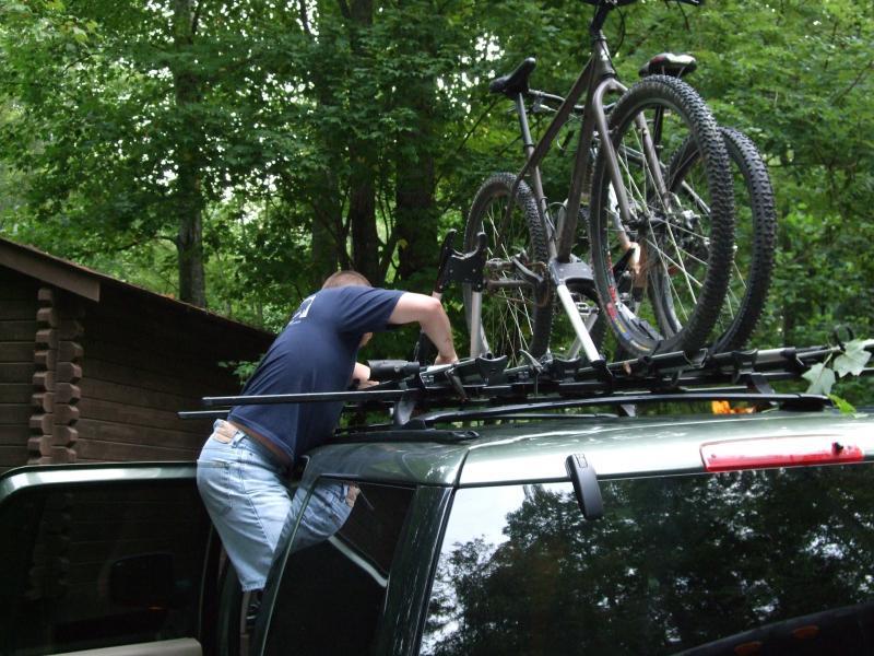 A person is loading a mountain bike onto the roof rack of a green SUV in a wooded area. The individual is partially leaning on the vehicle while securing the bicycle, surrounded by trees and a cabin in the background. Laurel Mountain mountain bike trail.