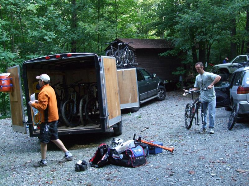 Two men are preparing for an outdoor activity in a wooded area. One man is unloading items from a trailer while the other is carrying a bike. The trailer is open, revealing several bicycles inside, and there are various sports bags and equipment on the ground. A parked vehicle is also visible in the background, along with a small cabin. Laurel Mountain mountain bike trail.