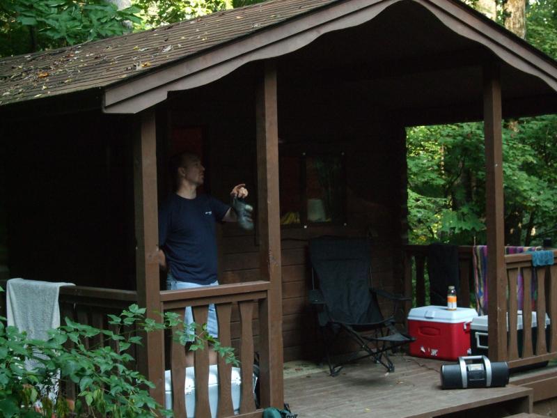 A man standing on the porch of a wooden cabin in a forested area, holding a small object in his hand. The cabin features a wooden railing and has outdoor seating. In the background, there are chairs, a cooler, and a couple of towels hanging. Lush greenery surrounds the scene, suggesting a peaceful outdoor setting. Laurel Mountain mountain bike trail.