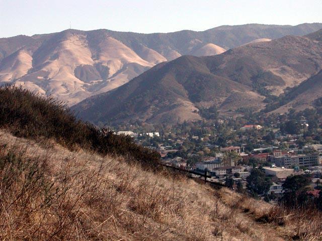 A panoramic view of rolling hills and mountains, with dried grassy slopes in the foreground and a valley with scattered trees and buildings below. The landscape features a mix of brown and green tones, illustrating a semi-arid environment. Madonna Mountain: Rock Garden mountain bike trail.