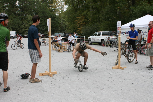 A group of people watching a bicycle competition in a gravel area, where a cyclist is attempting to ride under a low bar while balancing on a small bike. Some participants are cheering, and there's a tent in the background with bicycles and equipment.