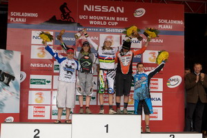 A podium ceremony at the UCI Mountain Bike World Cup, featuring five participants celebrating their achievements. The first, second, and third place winners are holding trophies and flowers, standing on the podium, with a backdrop displaying sponsor logos. An audience can be seen in the background applauding.