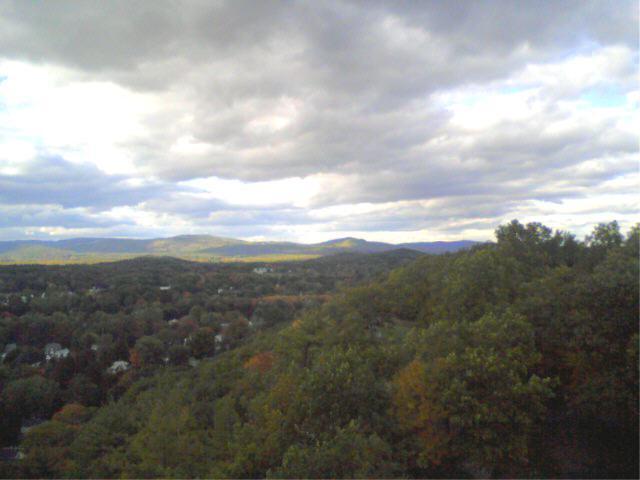 A scenic view of rolling hills and valleys, covered in trees with a mix of green and autumn colors. The sky is partly cloudy, with patches of blue peeking through. Small houses are visible in the valleys below, creating a serene landscape. Rocky Mountain And Highland Park mountain bike trail.