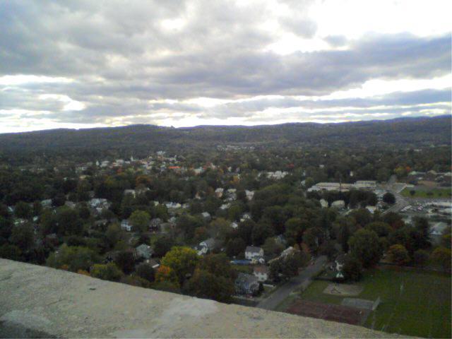 A panoramic view of a suburban landscape with a mix of residential homes and green trees under a cloudy sky. The scene includes rolling hills in the background and a hint of fall foliage. Rocky Mountain And Highland Park mountain bike trail.