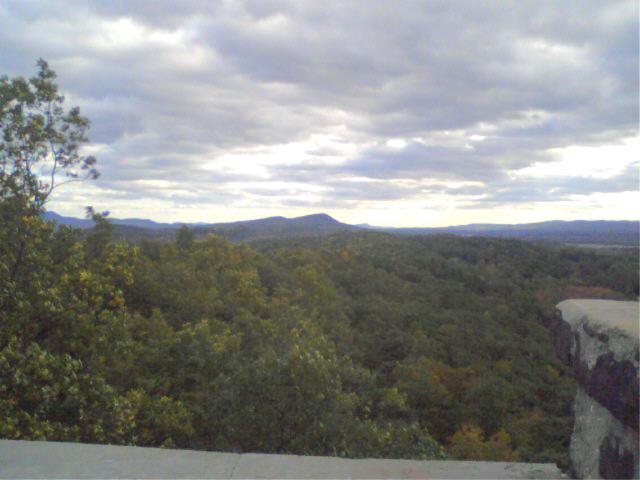 A panoramic view of a lush, green landscape from a rocky overlook, featuring rolling hills and a cloudy sky. The foreground includes vibrant tree foliage, and the distant mountains create a serene backdrop. Rocky Mountain And Highland Park mountain bike trail.