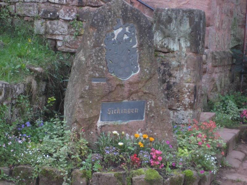 A weathered stone monument surrounded by colorful flowers, featuring an engraved coat of arms at the top and the name "Sickingen" on a metal plaque. The background includes lush green grass and stone structures. Landstuhl Trail mountain bike trail.