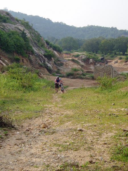 A cyclist walking a mountain bike along a rocky trail in a green, hilly landscape, with scattered boulders and trees in the background. The scene conveys a sense of outdoor adventure and exploration. Phnom Baset mountain bike trail.