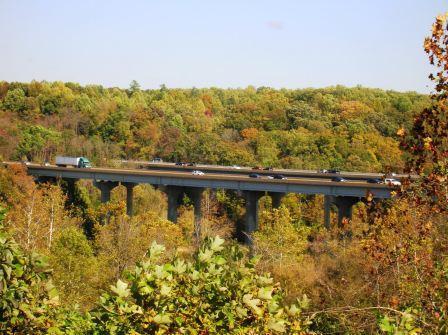 A scenic view of a highway bridge spanning over a forested area, with cars and trucks traveling along the road. The landscape features a rich tapestry of green and autumn foliage, showcasing a range of colors in the trees under a clear blue sky. Rappahanock River Trail mountain bike trail.