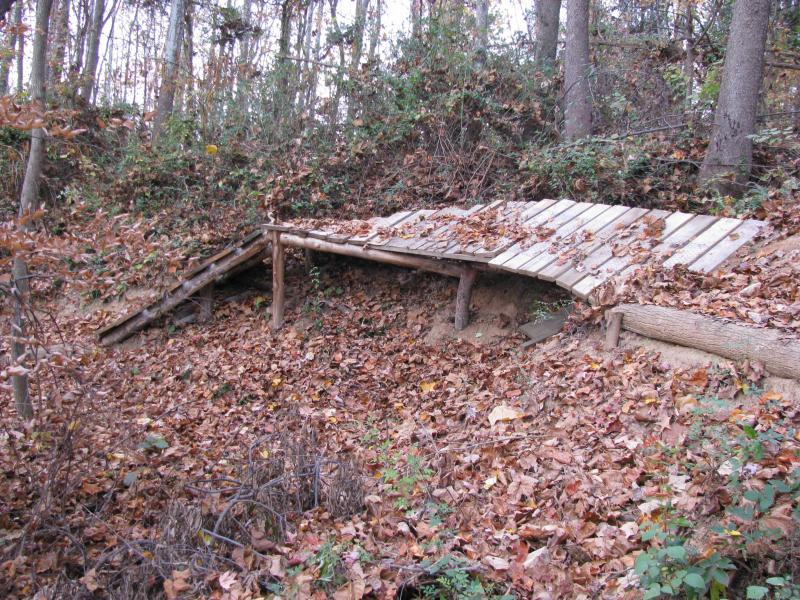 A wooden bridge spanning a small ravine covered with fallen leaves, surrounded by trees and underbrush. The bridge consists of planks resting on wooden supports, with earthy slopes leading to the sides. Rappahanock River Trail mountain bike trail.