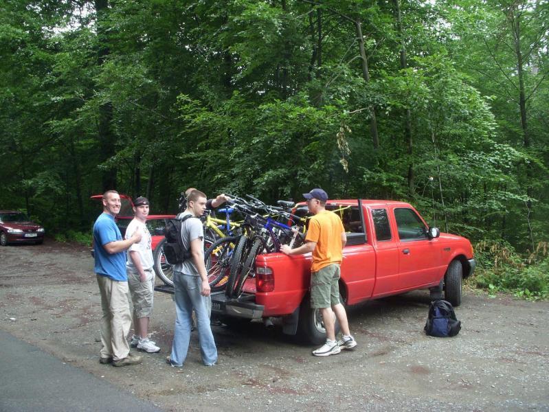 A group of five young men gathers around a red pickup truck parked in a wooded area. The truck's bed is loaded with various bicycles. Two of the men are securing the bikes, while the others are chatting and preparing for their outdoor activity. The scene is set in a natural environment, with trees and greenery surrounding them. Stempleberg mountain bike trail.
