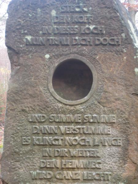Stone monument with an inscription in German. The top part challenges the reader to put their head through a circular opening, while the lower section contains poetic verses about understanding and making one's way easier. The surrounding environment features trees, suggesting a natural setting. Stempleberg mountain bike trail.