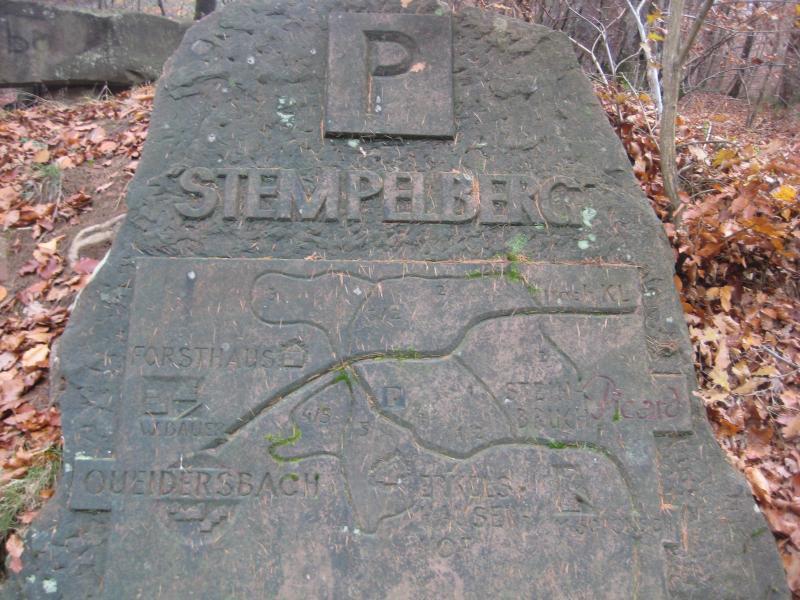 A weathered stone marker featuring the inscription "P STEMPBERG" at the top, with a map outline showing nearby locations such as Forsthaus and Queidersbach. The stone is surrounded by fallen leaves and forest scenery. Stempleberg mountain bike trail.