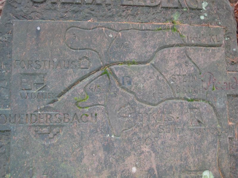 A weathered stone map displaying a trail or area, with inscriptions indicating locations such as "Forsthaus," "W. Bauer," and "Weidersbach." The surface has natural elements like moss and pine needles, suggesting it is located outdoors in a forested area. Stempleberg mountain bike trail.