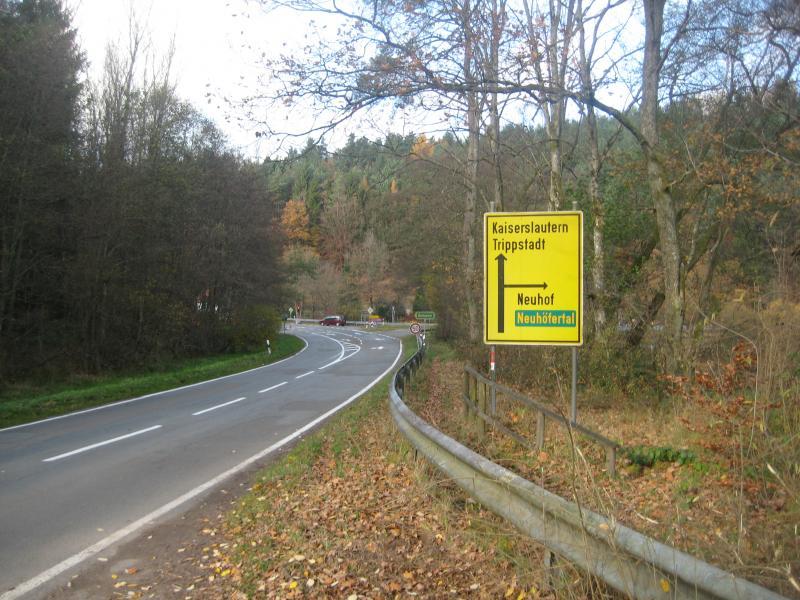 A yellow road sign indicating directions to Kaiserslautern and Trippstadt, with an arrow pointing left towards Neuhof and Neuhöfertal. The sign is positioned beside a winding road surrounded by trees in autumn foliage. Schopp Trail #3 mountain bike trail.
