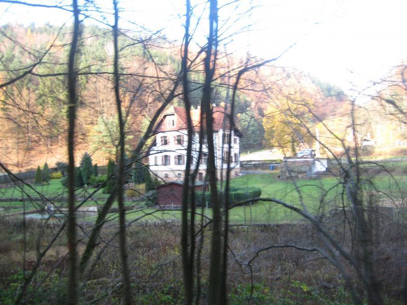 A large, multi-story house with a red-tiled roof, surrounded by lush greenery and trees. The house is partially visible through a cluster of bare branches in the foreground, with a serene landscape featuring a manicured lawn and nearby structures in the background. Schopp Trail #3 mountain bike trail.