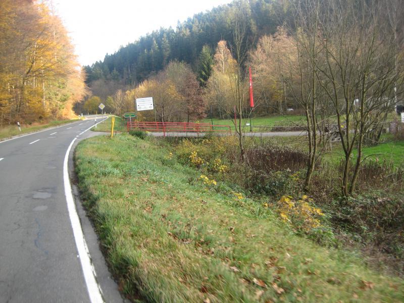 A winding rural road surrounded by trees in autumn colors, with a grassy shoulder and a small section of land with a red fence visible in the background. Road signs can be seen along the roadside, indicating directions and points of interest. Schopp Trail #3 mountain bike trail.