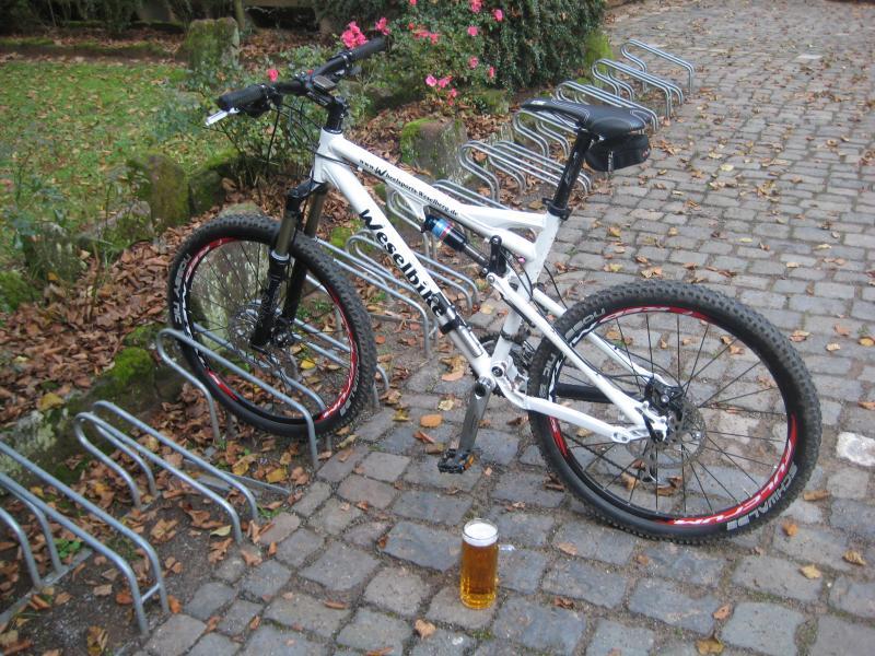 A white mountain bike parked on a cobblestone path, with a glass of beer placed nearby. Surrounding the bike are fallen leaves, and visible bike racks are in the background. Green foliage and shrubs can also be seen, indicating a natural setting. Schopp Trail #3 mountain bike trail.