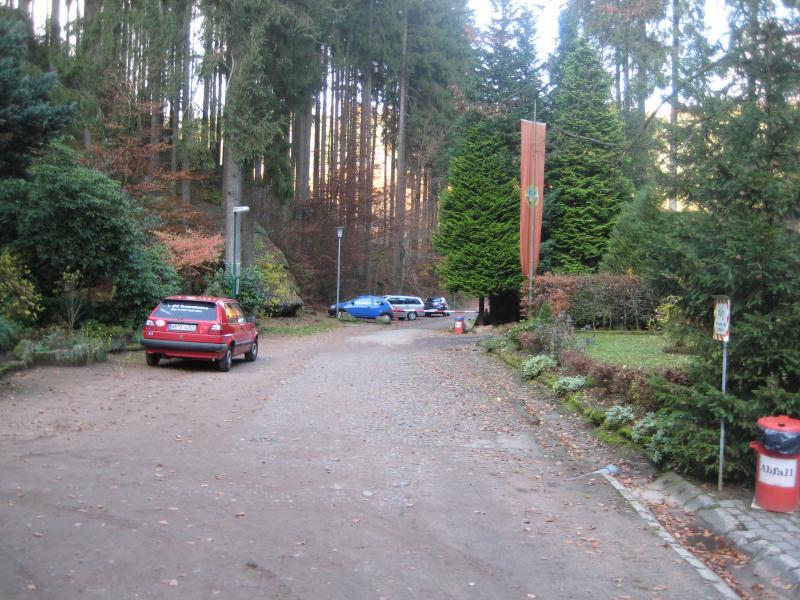 A scenic view of a gravel road lined with trees, leading into a wooded area. On the left, a red car is parked, and in the background, several other cars are visible among greenery. A flag hangs from a nearby tree, and a trash can marked "Abfall" is positioned on the right side of the path. The setting suggests a tranquil outdoor environment surrounded by nature. Schopp Trail #3 mountain bike trail.