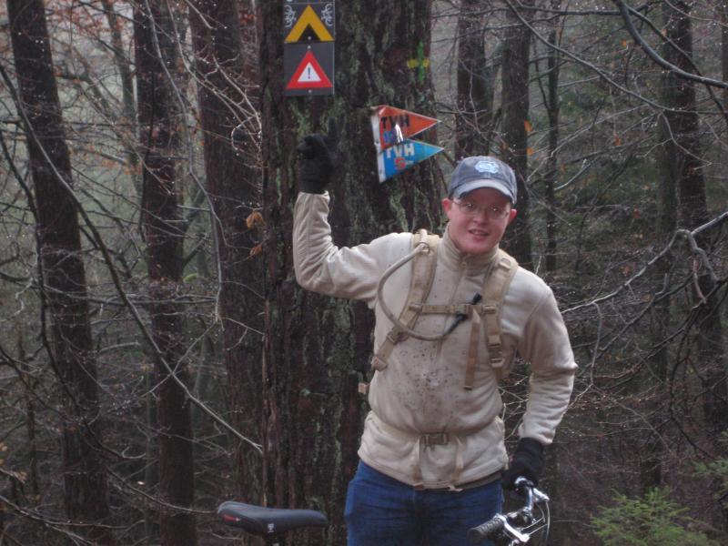 A smiling person wearing a beige long-sleeve shirt and a cap stands next to a tree in a wooded area, pointing to trail signs that indicate biking routes. The person is equipped with a backpack and is beside a mountain bike on the ground. The surroundings feature bare trees and a misty atmosphere. Schopp Trail #3 mountain bike trail.