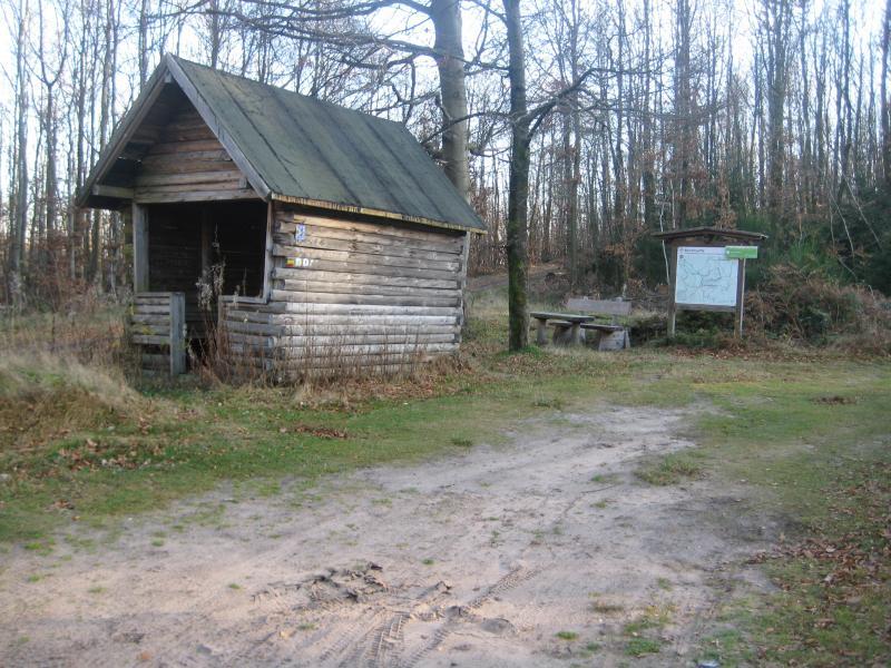 A rustic wooden shelter nestled in a forest clearing, with a worn path leading to it. Next to the shelter, there is a wooden table and a signboard with a map. The surrounding area features sparse grass and trees in a natural landscape, indicating a quiet outdoor setting. Johanniskreuz Trail 9 mountain bike trail.
