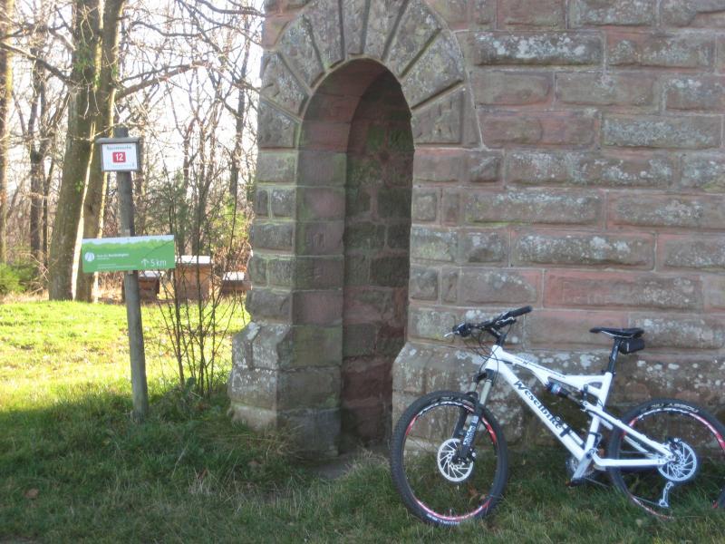 A mountain bike leaning against a stone structure with an arched doorway, next to two signs: one indicating a trail marker number 12 and the other showing a distance of 5 km. The scene is set in a grassy area with trees in the background and it appears to be a sunny day. Johanniskreuz Trail 9 mountain bike trail.