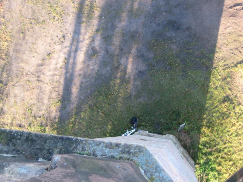 Aerial view of a grassy area with a paved path, taken from a high vantage point. The image shows shadows cast by surrounding trees and a small bicycle visible on the ground near the edge of a structure. Johanniskreuz Trail 9 mountain bike trail.