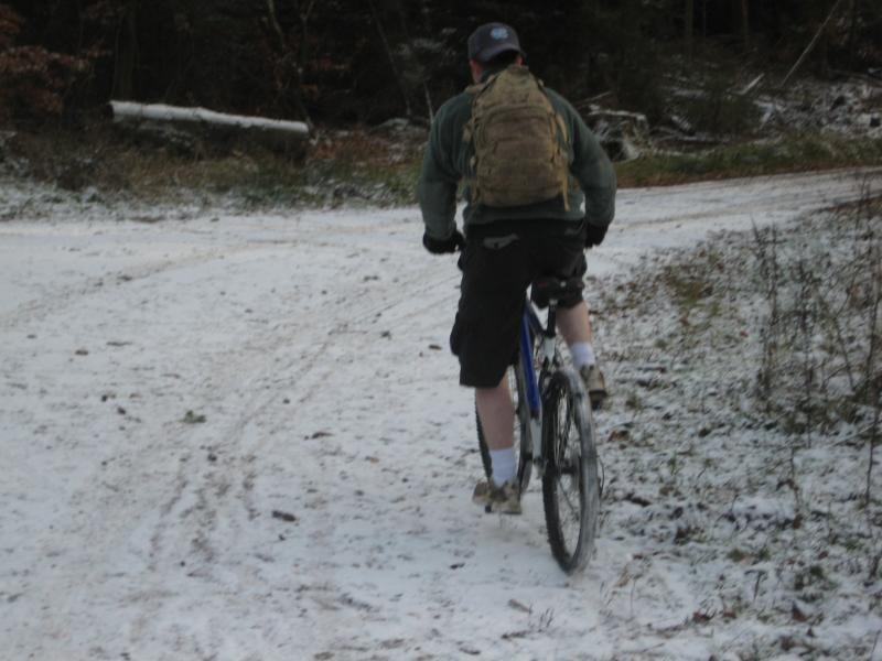 A person riding a mountain bike on a snowy path in a forested area. The cyclist is wearing a backpack and shorts, with visible snow covering the ground and surrounding foliage. The scene captures a sense of adventure and outdoor activity in winter conditions. Johanniskreuz Trail 9 mountain bike trail.
