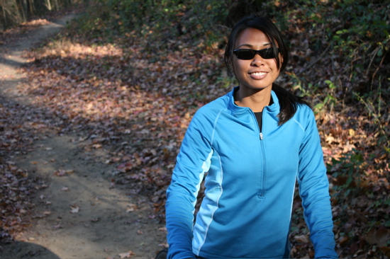 A smiling woman wearing sunglasses and a blue athletic jacket stands on a leaf-covered dirt trail in a forested area, with trees and foliage in the background.