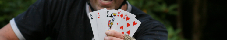 A close-up of a person smiling while holding a hand of playing cards, showing an Ace, King, and a pair of eights in hearts, with a blurred natural background.