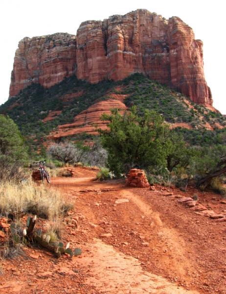 A scenic view of a red rock mountain towering over a dirt trail, surrounded by desert vegetation, including shrubs and cacti. The winding path leads towards the mountain base, showcasing the rugged landscape typical of a southwestern region. Bell Rock Trailway mountain bike trail.