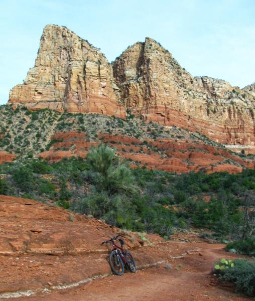 A mountain bike rests on a rocky trail in a vibrant desert landscape, featuring towering red rock formations and lush vegetation in the foreground. The sky is clear with a few scattered clouds, highlighting the rugged beauty of the terrain. Llama Trail mountain bike trail.