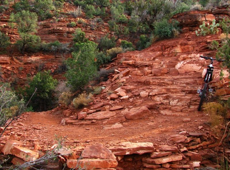 A rocky mountain biking trail winding through desert terrain, with vibrant green shrubs and trees scattered among red stone formations. A black mountain bike is parked on the trail. Templeton mountain bike trail.