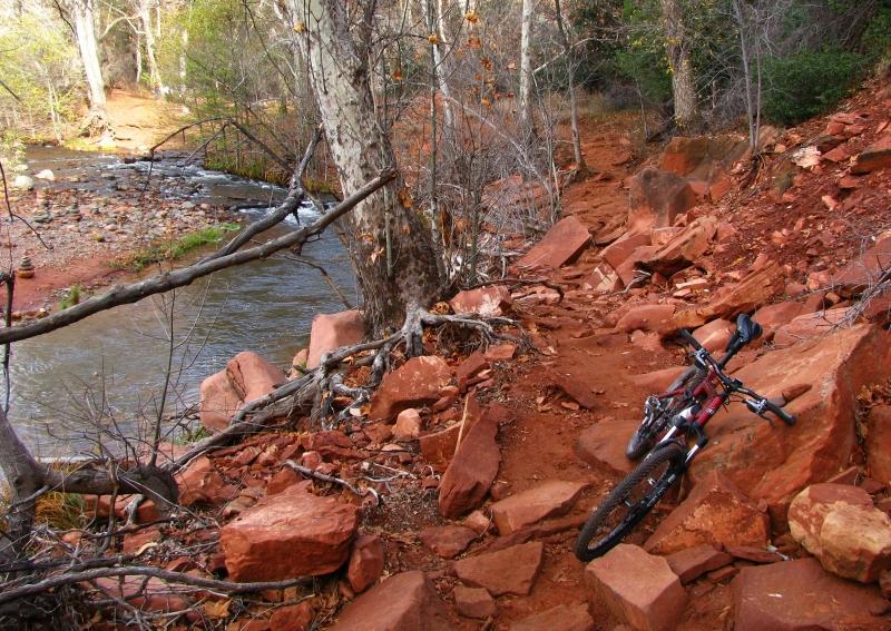 A mountain bike resting on a rocky, reddish dirt trail next to a flowing creek, surrounded by bare trees and greenery. Templeton mountain bike trail.