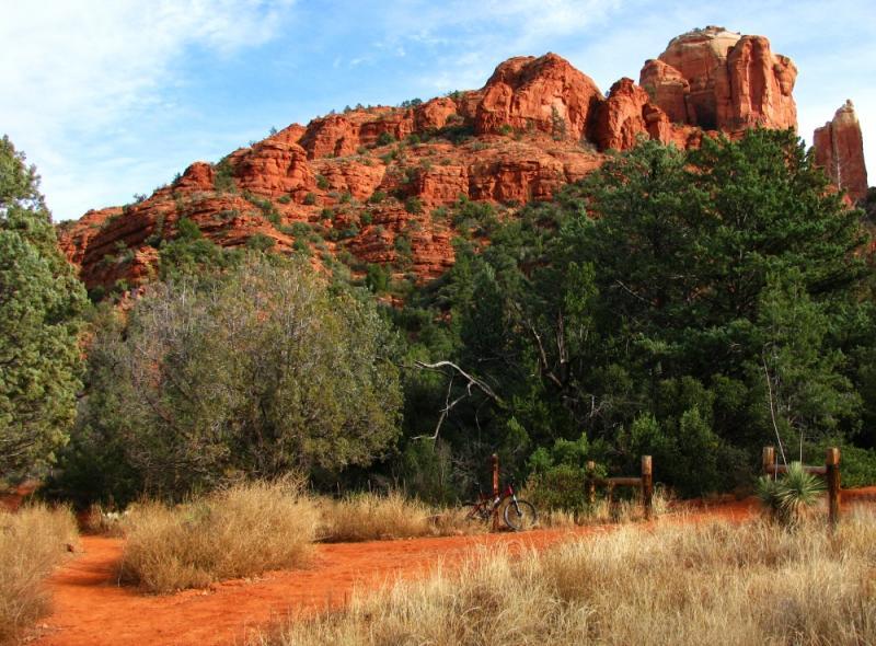 A scenic view of red rock formations in a wooded area, featuring green trees and dry grass. A bicycle leans against a fence along a winding dirt path that leads into the landscape. The sky is clear with some clouds, highlighting the vibrant colors of the surroundings. Templeton mountain bike trail.