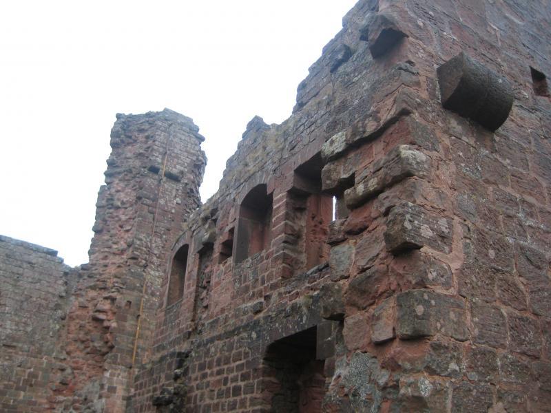 Ruins of an old stone structure, showcasing weathered red bricks and various openings for windows. The image captures a portion of the building at an angle, highlighting the texture of the stones and remnants of a tower in the background, against a cloudy sky. Hohenecken Trails mountain bike trail.