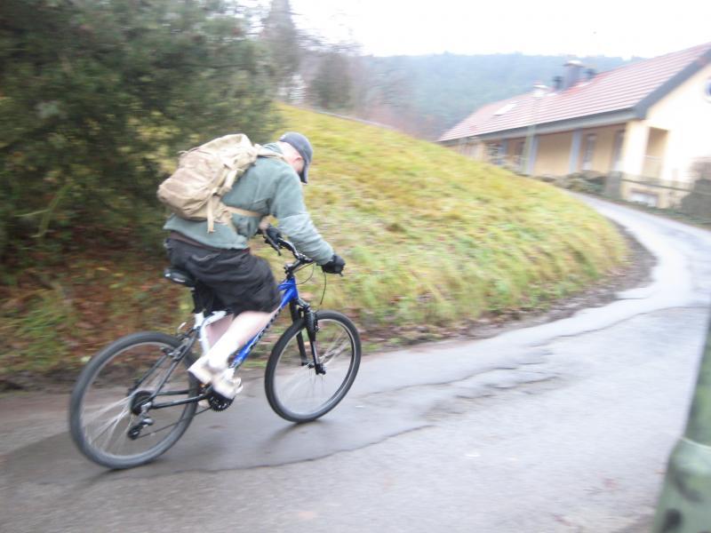 A person riding a blue mountain bike down a wet, winding road. The rider is wearing a green sweatshirt, shorts, and a backpack, with a hat on their head. Grass and trees are visible on the right side, along with a yellow house in the background. The scene appears to be overcast and possibly rainy. Hohenecken Trails mountain bike trail.